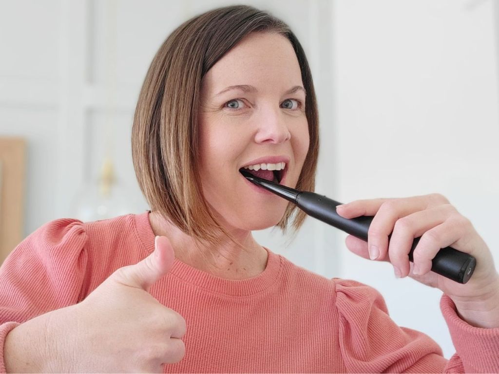 Woman using an electric toothbrush with her left and and giving a thumbs up with her right hand.