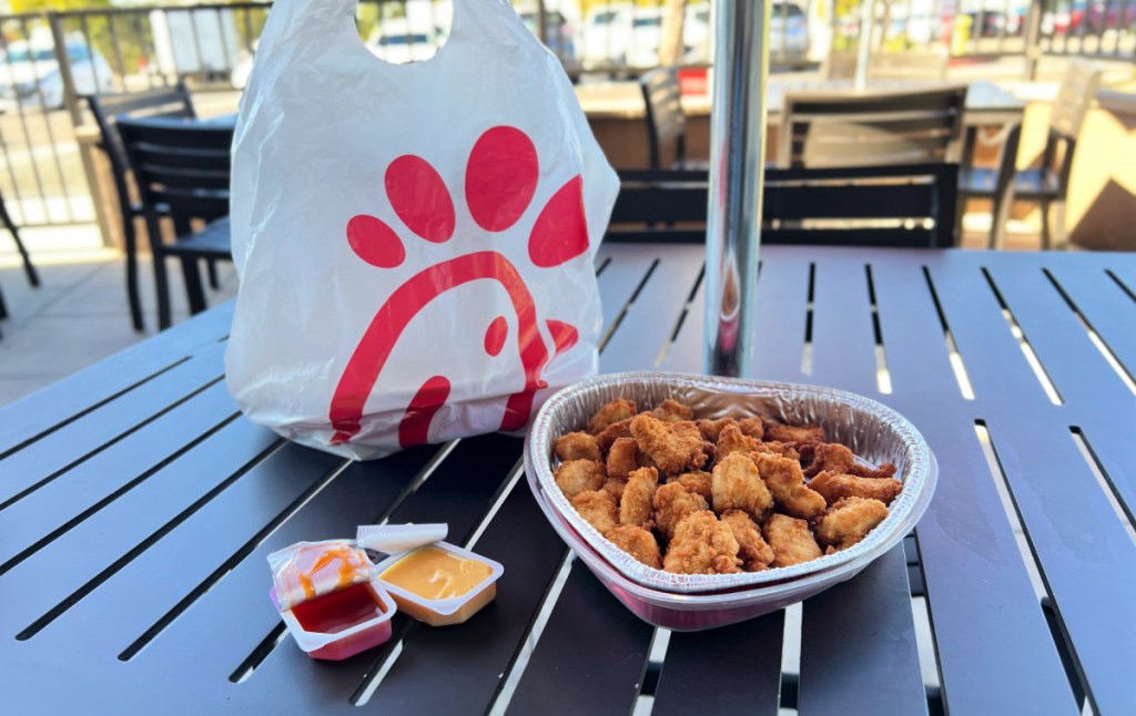 heart shaped tray of chicken nuggets next to a chick fil a togo bag and sauces under an outdoor dining table umbrella