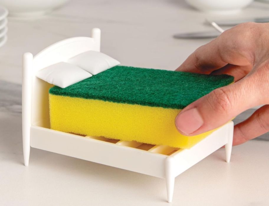 a hand grasping a sponge on a bed-shaped sponge holder with pillows