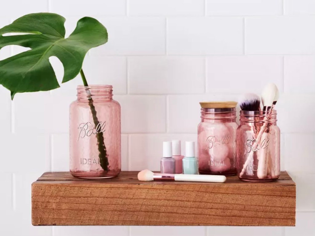 three pink vintage ball jars on shelf with makeup brushes and plant in them
