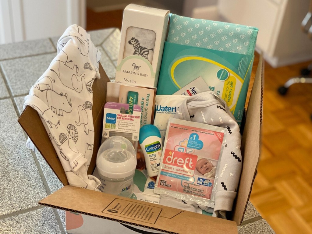 Box filled with baby items including Dreft sample, baby bottle, baby blanket, onesie, and Cetaphil sample.
