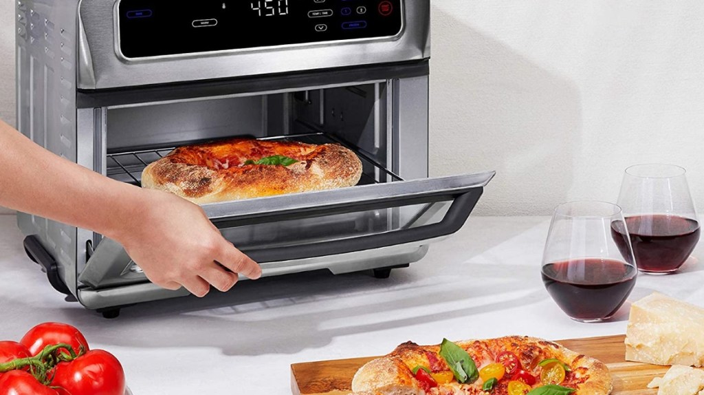 Hand opening an air fryer with a pizza inside. Two wine glasses and a pizza on a cutting board are sitting next to the air fryer.