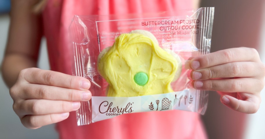 little girl holding up a flower shaped cookie with yellow frosting and a green candy in the center