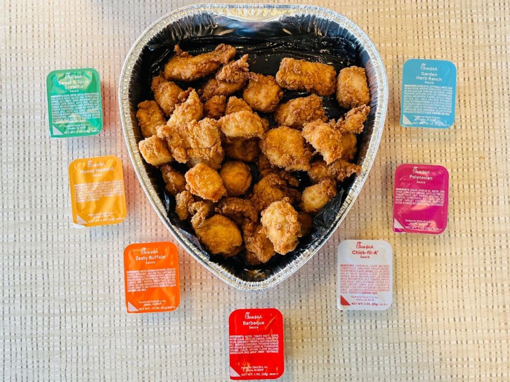 A heart shaped tray filled with Chick-fil-A nuggets with containers of different dipping sauces evenly spaced out around the heart