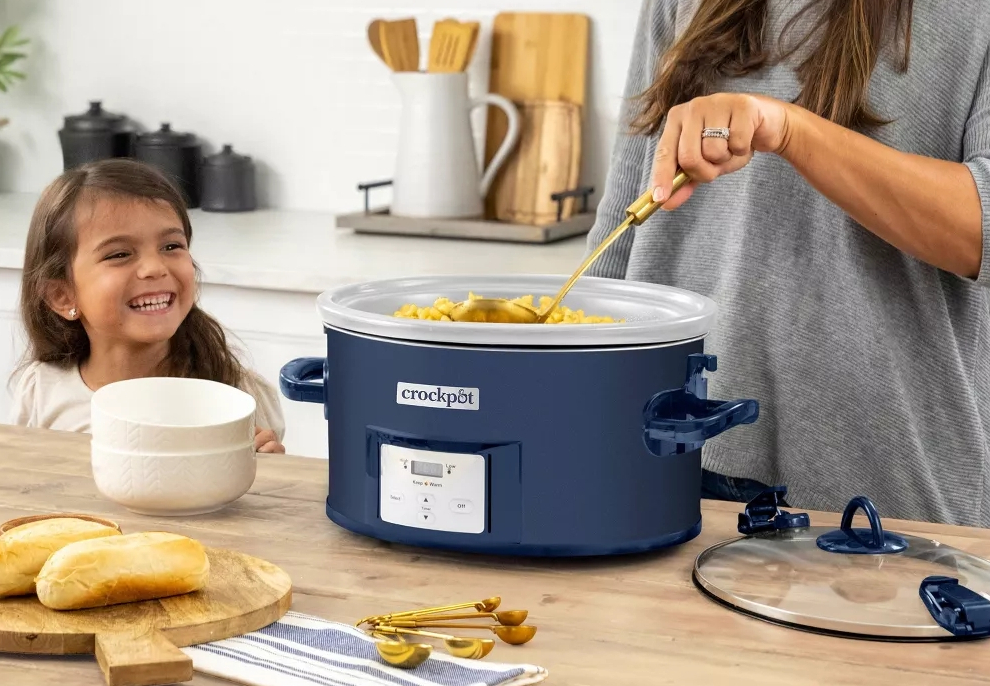 Woman stirring food in a slow cooker while a girl stands next to her smiling.
