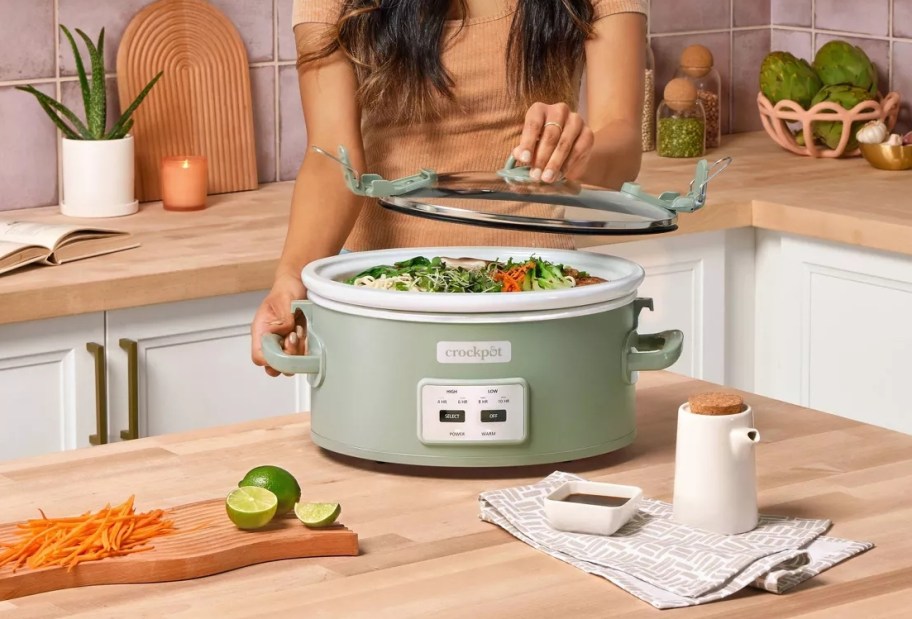 A woman is lifting the lid off of a Crockpot and checking the food inside. The Crock Pot is sitting on a kitchen counter.