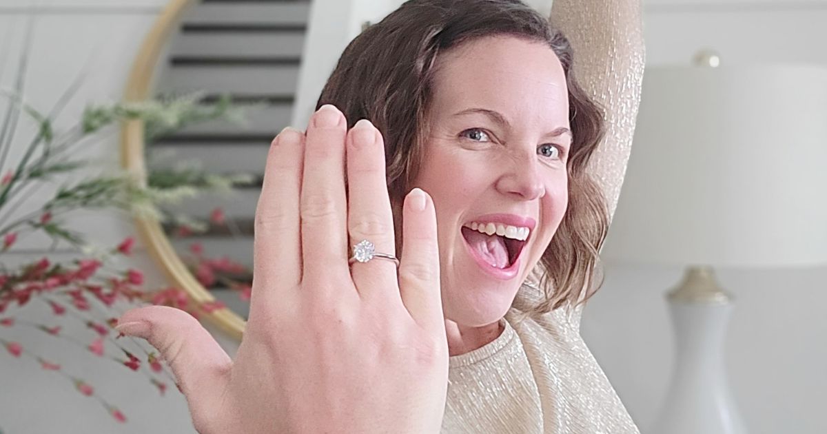 woman holding up her heand with an engagement type ring on her ring finger with a very excited expression on her face