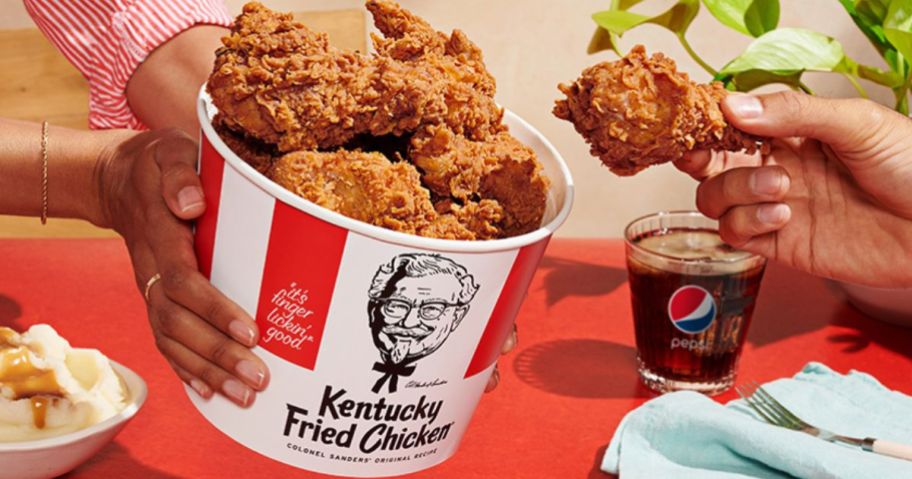 A hand taking a chicken drumstick out of a KFC bucket of chicken
