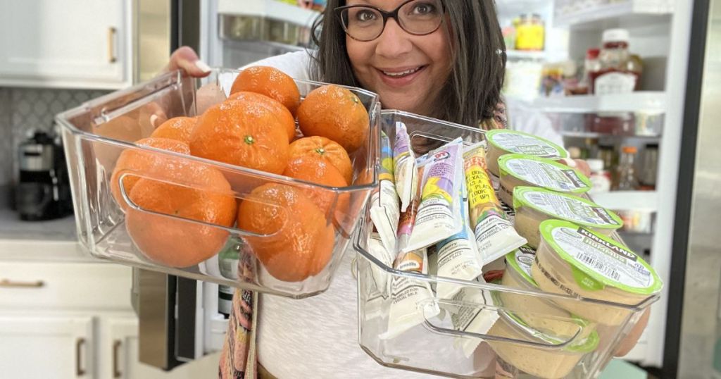 woman holding clear bin filled with oranges in one hand and clear bin filled with snacks in the other hand