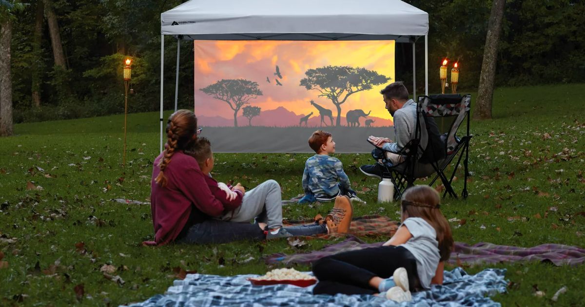 Ozark trail shade projector screen attached to support poles of a 10ft x 10ft canopy with a family seated around watching a movie