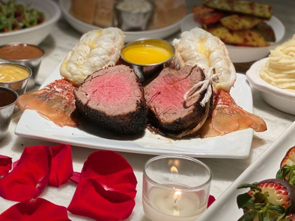 Filet Mignon and lobster tail dinner surrounded by side dishes, roase petals and a lit votive candle in a glass candle holder
