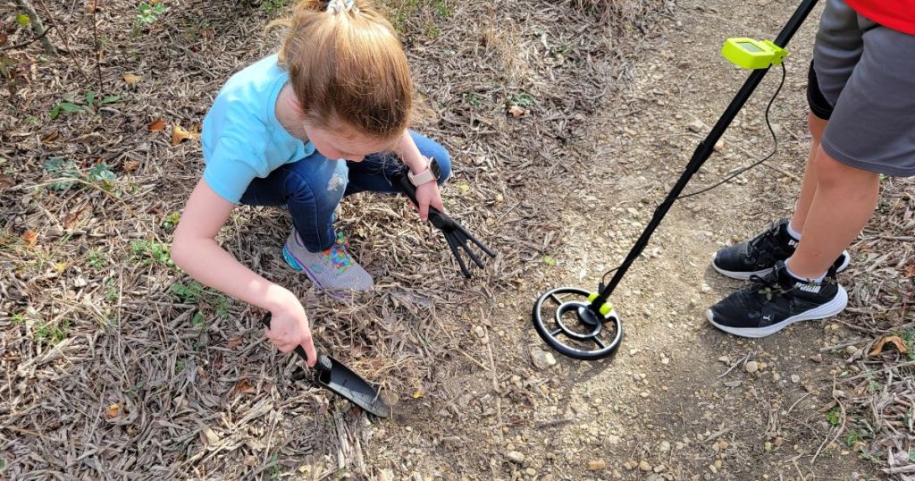 kid using metal detector while girl is starting to dig into ground with hand rake and hand shovel