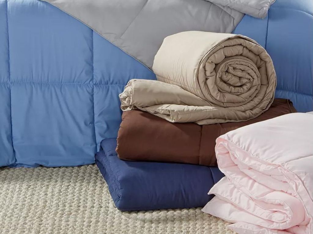various colors of comforters folded and rolled up on the floor by the bed