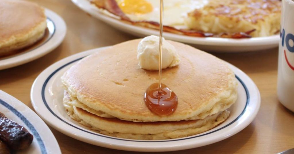 A stack of pancakes topped with a butter ball with syrup being drizzled on