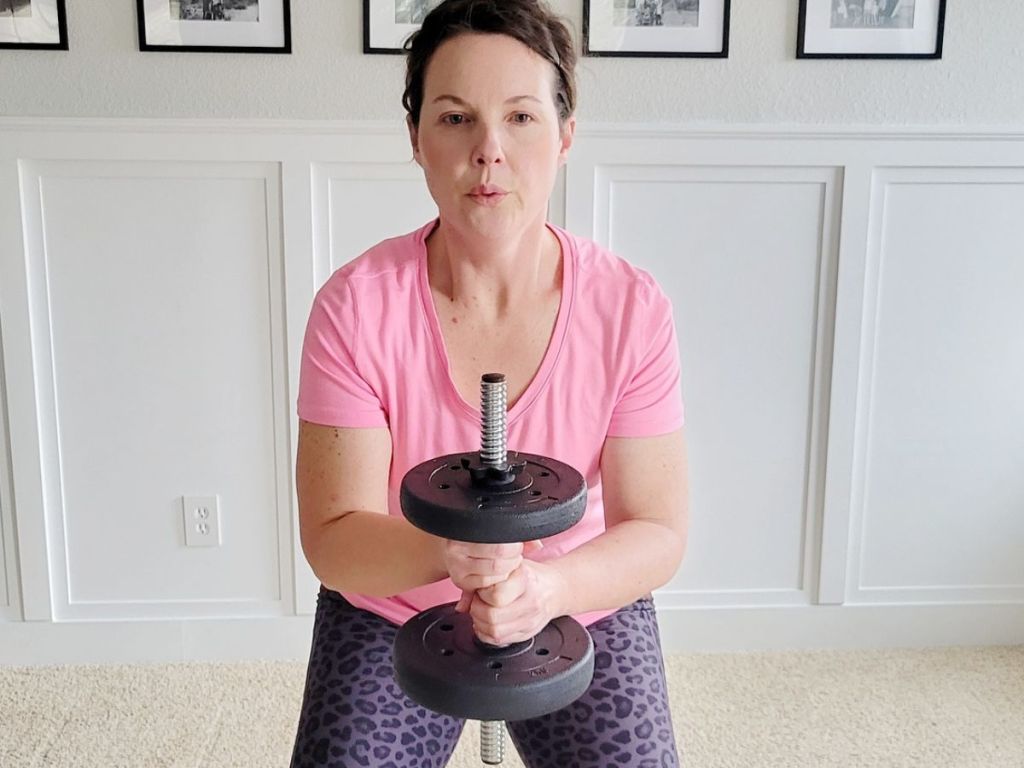 woman holding a heavy dumbell vertically in front of her with both hands while doing squats in her living room