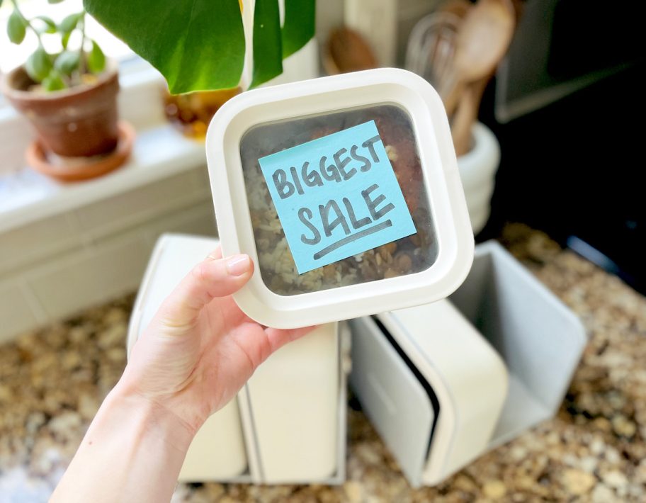 Hand holding a food storage container with blue post it note biggest sale on front 