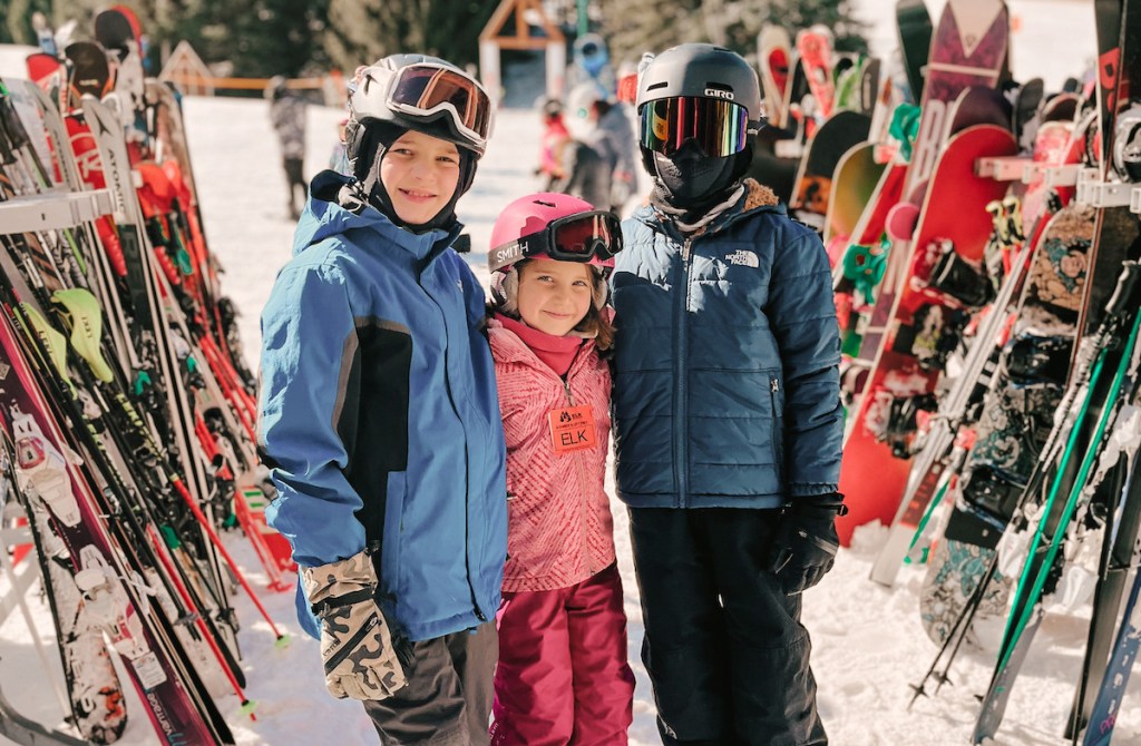 kids wearing ski gear in front of racks with snowboards and skis at resort