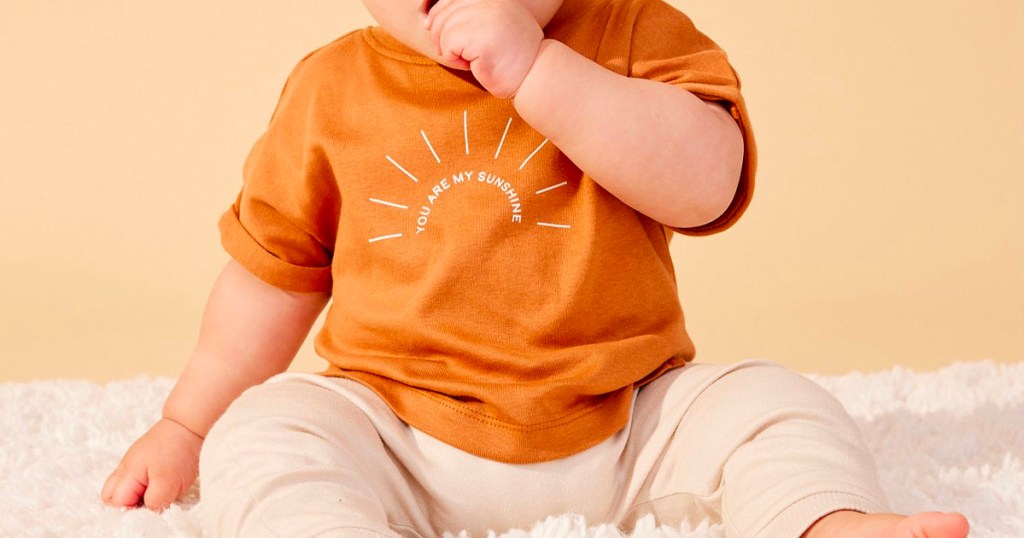 baby wearing little organics orange shirt and biege pants on carpet