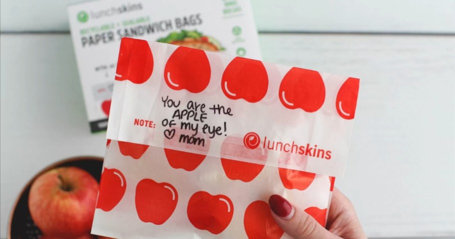 hand holding a paper sandwich bag with red apples on it and a note, the box they come in is in the background