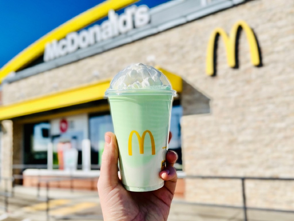 man's hand holding up a mcdonald's shamrock shake in front of a mcdonald's restaurant. 