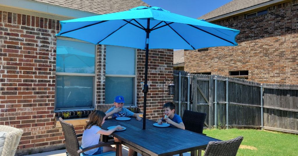 3 kids eating on patio table under blue umbrella