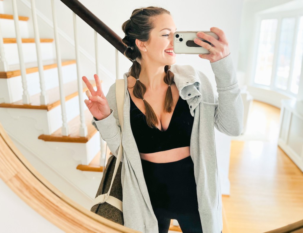 woman giving peace sign holding yoga mat and phone in mirror