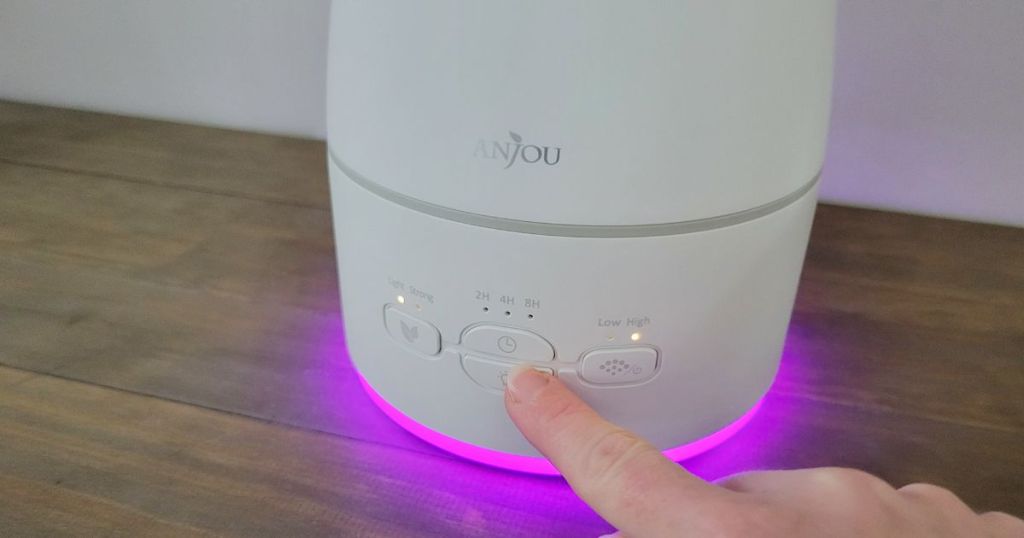 person pushing button on white Anjou diffuser with purple light placed on wood table 