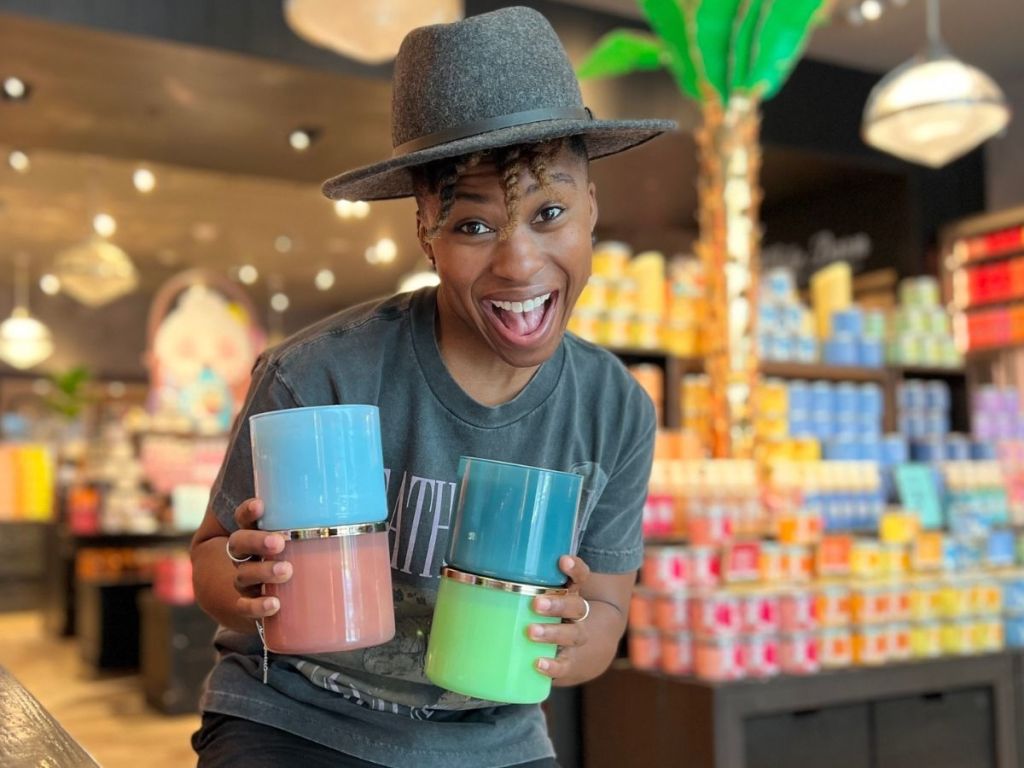Woman at bath & Body Works holding four 3-wick candles and looking very excited