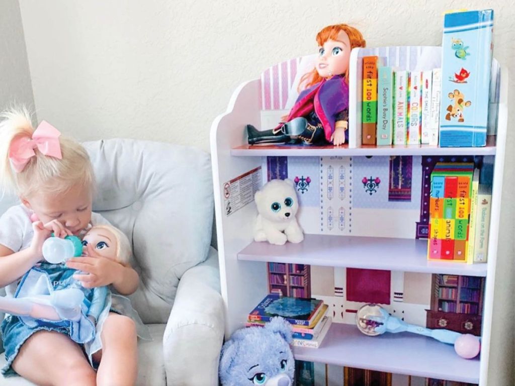 Small blonde child giving a bottle to a baby doll near a bookshelf