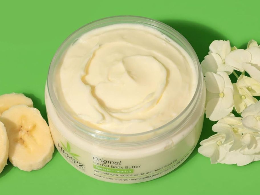 A jar of Hempz Body Butter next to banana slices and vanilla flower blossoms