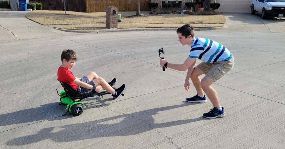 two children playing in the street, with one riding a go-cart type vehicle and the other filming on a phone using a gimbal