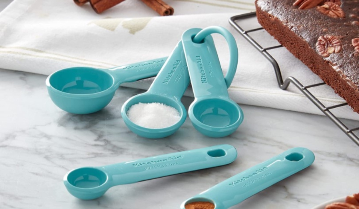 Kitchen Aid Measuring spoons on a countertop with a cake. One spoon has sugar in it and another has cinnamon in it.