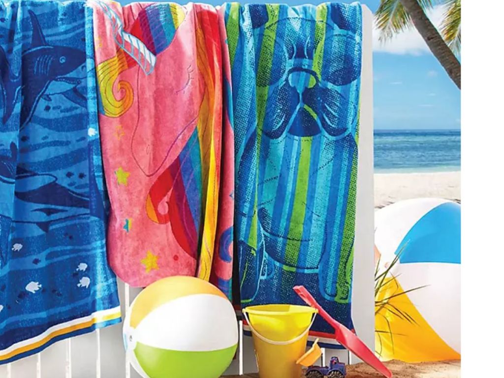 3 Member's mark Kids beach towels hanging on a fence outside with a beach ball, sand pail, and a shovel