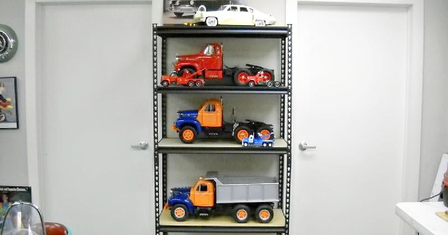 A black metal shelf displays model trucks and cars. The top shelf holds a white car, the middle shelves house red and orange trucks.