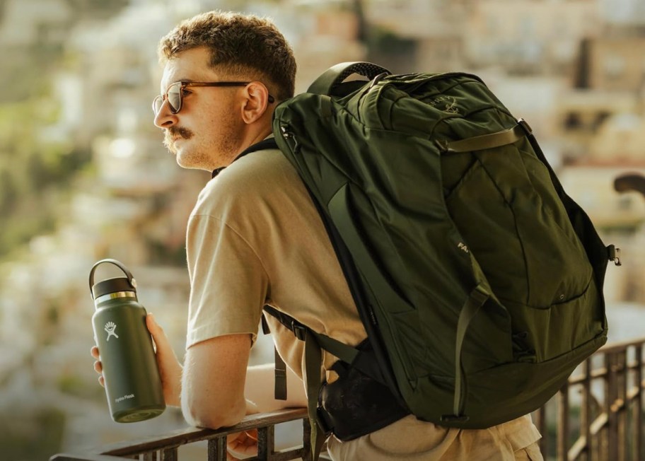man leaning over railing holding hydro flask and wearing large backpack