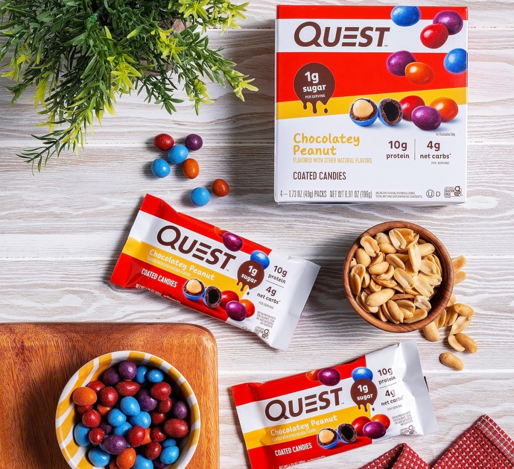 box and two bags of Quest Nutrition Chocolatey Peanut Coated Candies on table with bowl of candies and peanuts