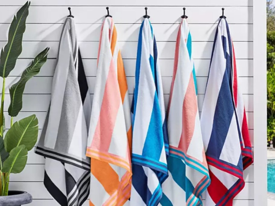 Different color, striped beach towels, hanging off black pegs with white fence in the background