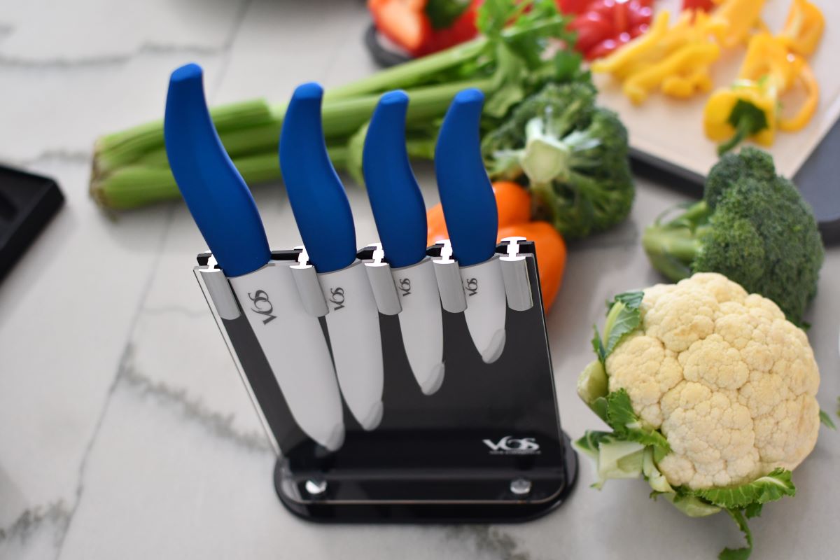 Four knives with blue handles in a holder on a counter with vegetables around it
