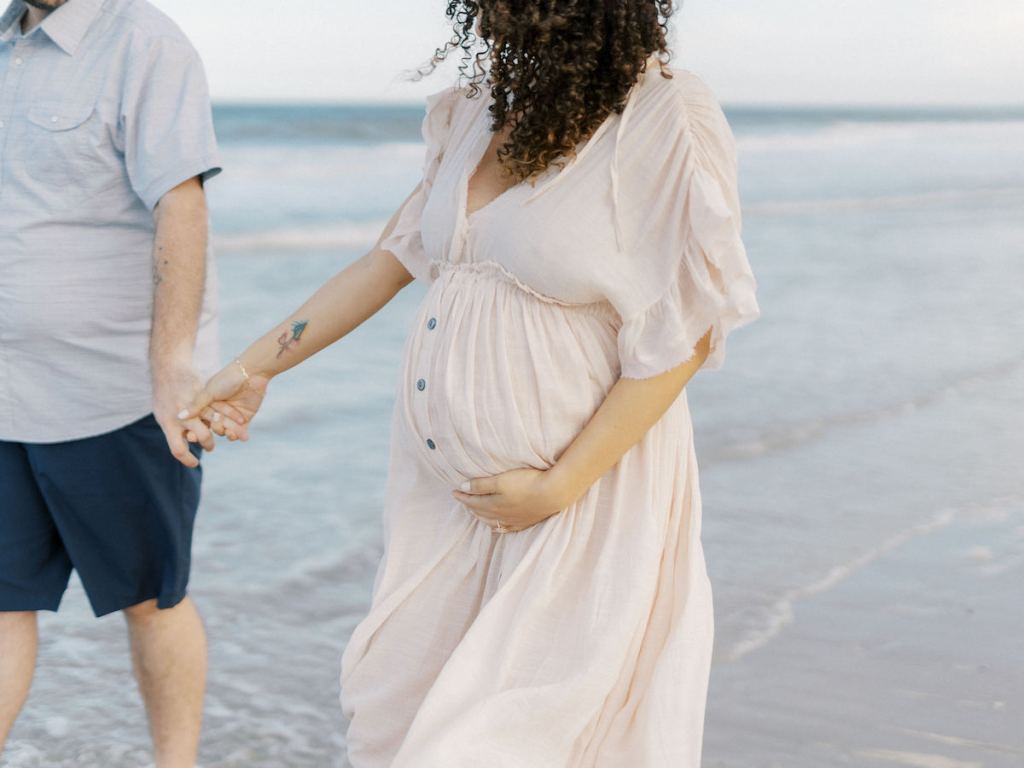 woman wearing blush colored button down dress holding bump and mans hand on beach