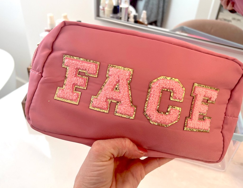 hand holding pink cosmetic bag with varsity patches that spell face