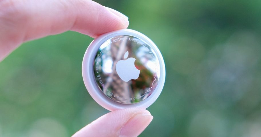 close up of fingers holding travel products apple airtag in front of greenery