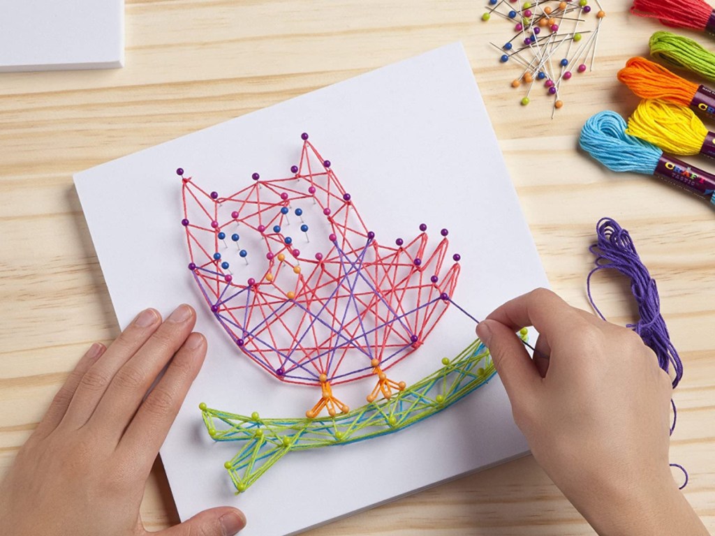 child's hands shown working on a craft-tastic string art owl kit next to bundles of colorful string and pins