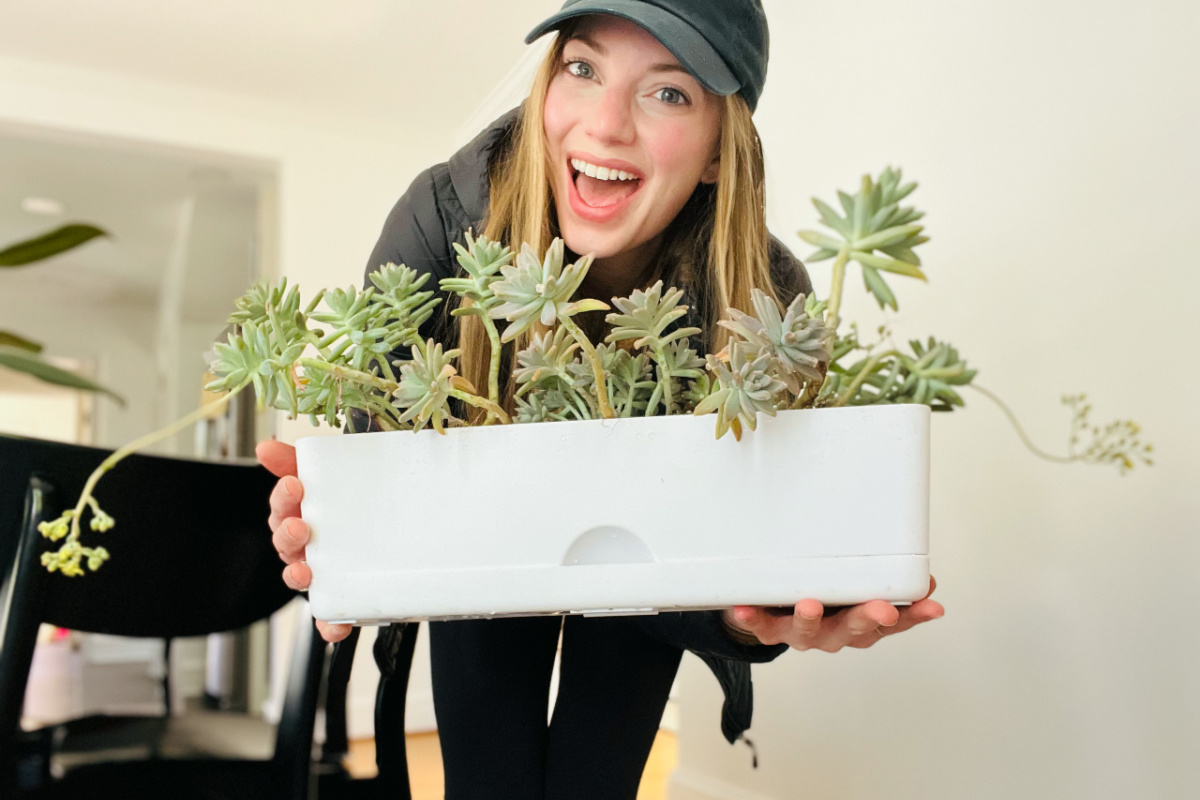 smiling woman in a baseball cap and jacket holding a white plastic self watering planter from target with plants in it