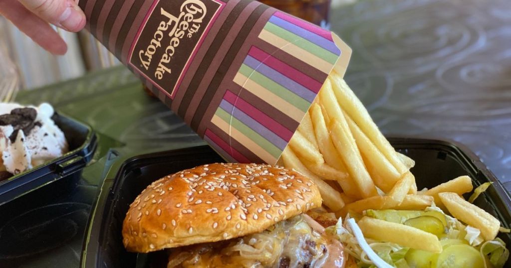 Fries being dumped into a cheesecake factory takeout with a burger