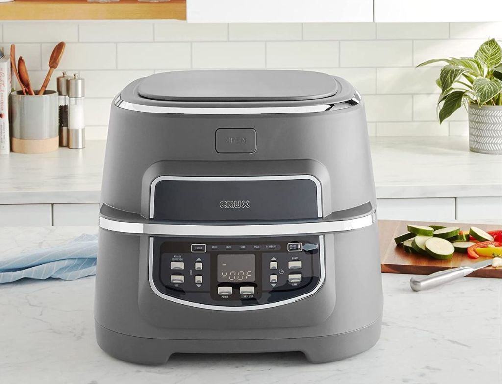 Silver air fryer sitting on a kitchen counter with a cutting board of food behind it