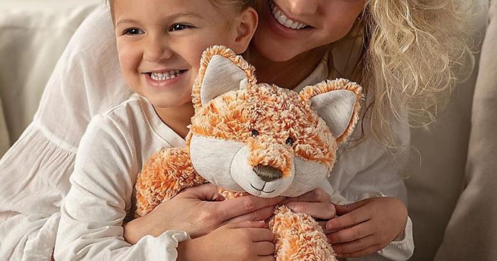 Little girl sitting in a woman's lap and holding a DEMDACO Heartful Hugs fox plush