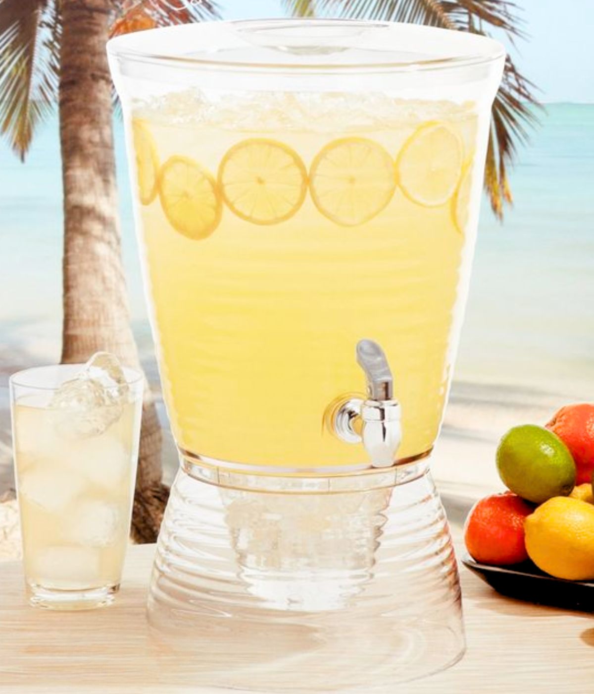 A 2.5 gallon drink dispenser filled with lemonade and ice sitting next to a glass of lemonade and a plate of citrus fruits