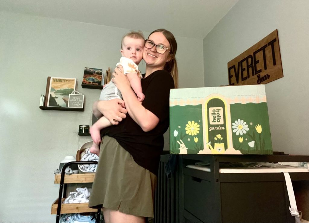 Woman holding a baby and standing next to a Hello Bello box