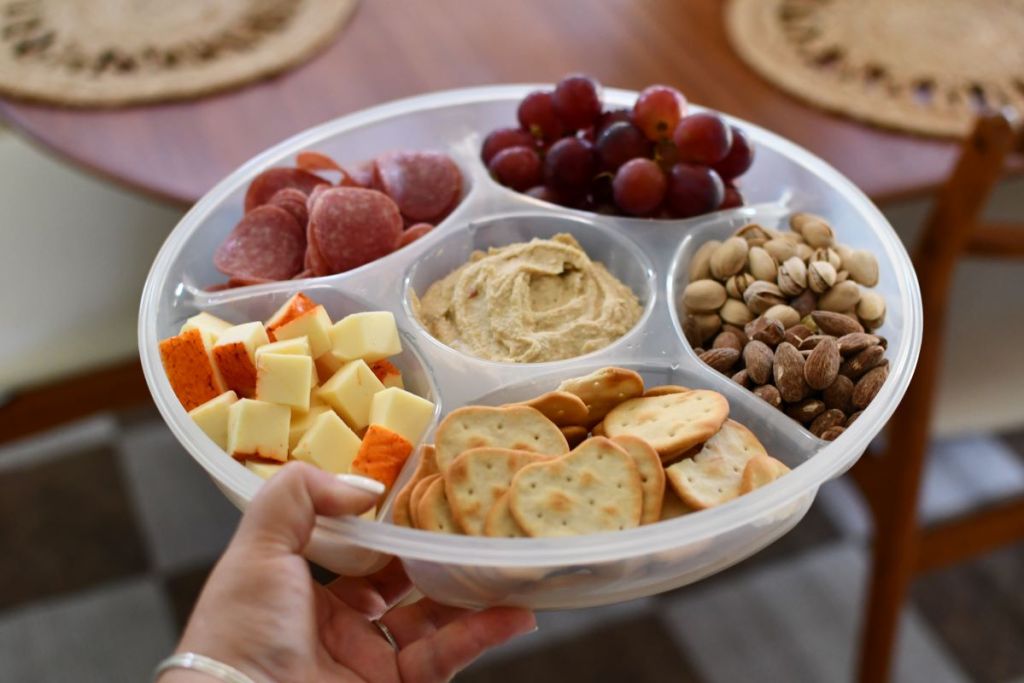 Hand holding a snack tray with cheese, crackers, nuts, fruit, and dip
