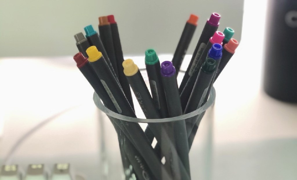 glass cup full of journaling pens in various colors sitting on a desk